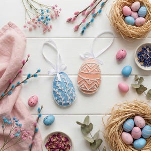 May include: Overhead shot of Easter decorations on a white wooden surface. Two decorative egg ornaments with white ribbon hang from the top. The eggs are blue and peach, with white bead detailing. Other decorations include painted eggs, nests, and flowers.