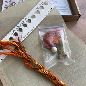 May include: A cross-stitch kit featuring a beige linen fabric, a white thread organizer labeled Tela Artis with Support Ukraine text, a braided bundle of orange, yellow, and green embroidery floss, and a small plastic bag containing a heart-shaped charm.