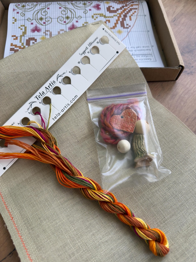 May include: A cross-stitch kit featuring a beige linen fabric, a white thread organizer labeled Tela Artis with Support Ukraine text, a braided bundle of orange, yellow, and green embroidery floss, and a small plastic bag containing a heart-shaped charm.