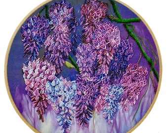 May include: A beaded floral embroidery in a wooden hoop. The artwork features cascading wisteria blossoms in shades of purple, lavender, and white, with green beaded vines. The background is a gradient of purple hues.