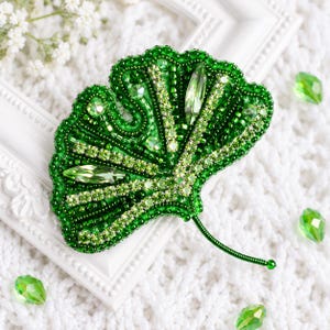 May include: A ginkgo leaf brooch crafted with vibrant green beads, sparkling rhinestones, and faceted crystals. The design features a fan-shaped structure with a thin, wire-wrapped stem, showcasing intricate beadwork and varied shades of green.