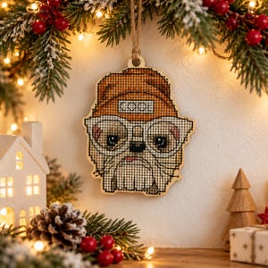 May include: A wooden cross-stitch ornament shaped like a dog wearing an orange beanie with the word COOL printed on it. The dog has large glasses, a small tongue sticking out, and is suspended by a brown twine string for hanging.