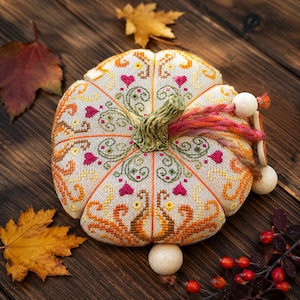 Finished embroidered pumpkin pincushion displayed on a rustic wooden tabletop surrounded by autumn leaves and berries. Decorative stitches, hand-dyed floss, ceramic embellishments, and sampler-inspired motifs create a warm seasonal design perfect for fall decorating.