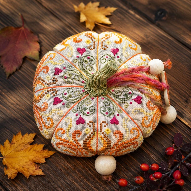 Finished embroidered pumpkin pincushion displayed on a rustic wooden tabletop surrounded by autumn leaves and berries. Decorative stitches, hand-dyed floss, ceramic embellishments, and sampler-inspired motifs create a warm seasonal design perfect for fall decorating.