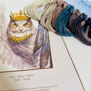 Puede incluir: Un kit de punto de cruz con un dise&ntilde;o de b&uacute;ho coronado. Incluye hilos de bordar de varios colores y tela blanca. El embalaje muestra el texto "Owl * Hibou * B&uacute;ho * Eule * Пугач" y mide 26,5 x 42,5 cm.