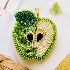May include: A handcrafted apple brooch featuring intricate beadwork in shades of lime, emerald, and forest green. The design incorporates sparkling crystals, a dark green beaded leaf, and a central section of cream-coloured seed beads with black accents.