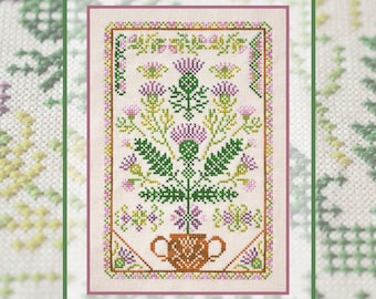 May include: A cross-stitch embroidery on light beige fabric featuring a central thistle plant with purple flower heads and green leaves, set in a brown two-handled pot. The design is enclosed within a decorative geometric border in pink and green.