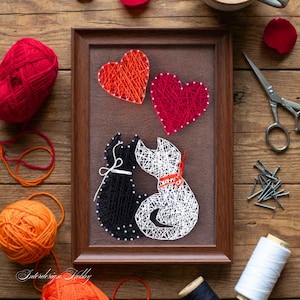 Puede incluir: Una pieza de arte de hilo enmarcada con dos gatos y dos corazones. Un gato es negro con un lazo blanco, el otro es blanco con un collar naranja. Los corazones son rojos y naranjas. La obra de arte está sobre un fondo marrón.