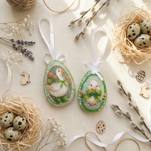 May include: Two embroidered Easter egg ornaments with white ribbon hangers. One features a goose, the other a duckling, both surrounded by floral designs. The eggs are light green with beaded edges, set among nests, eggs, and sprigs.