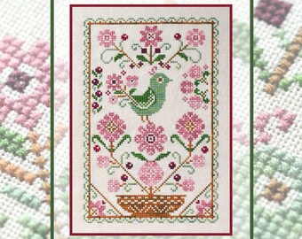 May include: A cross-stitch embroidery on white fabric featuring a central green bird perched above a brown woven basket. The design includes pink flowers, green stems, and small dark red accents, all enclosed within a decorative green and red border.