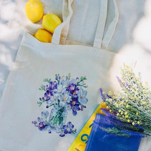 May include: A cream-colored tote bag with a floral design featuring purple and white flowers in a glass jar. The bag contains three yellow lemons. A blue notebook and a yellow magazine are next to the bag.