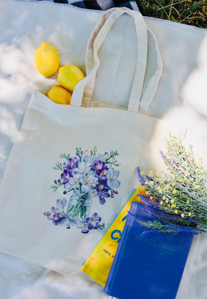 May include: A cream-colored tote bag with a floral design featuring purple and white flowers in a glass jar. The bag contains three yellow lemons. A blue notebook and a yellow magazine are next to the bag.