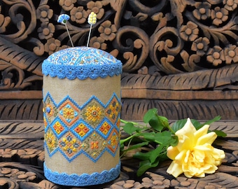 May include: A cylindrical pin cushion featuring a tan linen body with a blue and orange geometric cross-stitch pattern. The top and bottom edges are trimmed with blue crochet lace. Two decorative pins with small floral toppers are inserted.