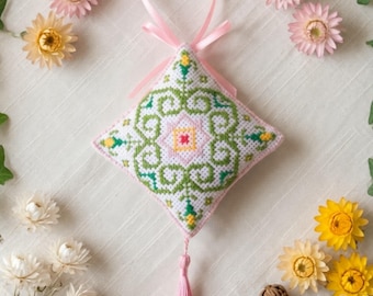 以下が含まれることがあります： 白い布地に緑と黄色の対称的な花柄が刺繍された、ひし形のクロスステッチオーナメント。上部には淡いピンクのリボンループ、下部の先端には同色のピンクのタッセルが取り付けられています。