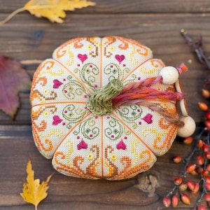 Top view of the finished pumpkin pincushion displaying symmetrical decorative stitching and ornamental sampler motifs. The dimensional pumpkin shape is accented by a ceramic stem, hand-dyed decorative thread, wooden beads, and vibrant autumn colors inspired by the fall season.