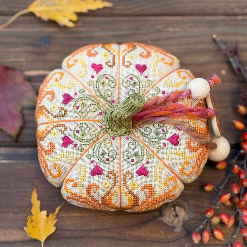 Top view of the finished pumpkin pincushion displaying symmetrical decorative stitching and ornamental sampler motifs. The dimensional pumpkin shape is accented by a ceramic stem, hand-dyed decorative thread, wooden beads, and vibrant autumn colors inspired by the fall season.