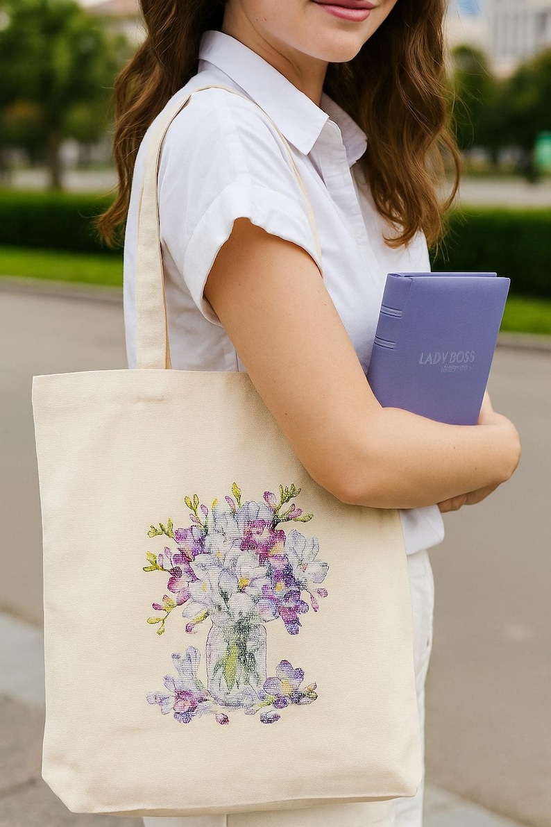 May include: Cream-colored tote bag featuring a floral design in shades of purple, white, and green. The bag is held by a person wearing a white shirt, along with a purple notebook labeled "LADYBOSS".
