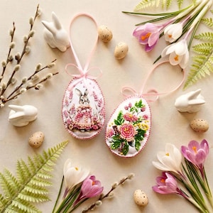 May include: Two decorative Easter egg ornaments with pink ribbons. One features a bunny in a pink dress, the other has a floral design. White ceramic bunnies, speckled eggs, and purple and white crocus flowers surround the eggs.