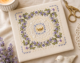 May include: A square piece of cream-coloured fabric featuring intricate cross-stitch embroidery of purple flowers and green vines. The design includes a central gold-toned winged heart charm and four small purple buttons at the corners of the floral border.