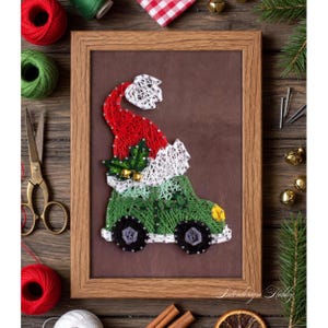 May include: A framed string art piece depicting a green car with a Santa hat. The artwork is made with red, white, and green thread on a brown background, set in a wooden frame. Christmas-themed decor.