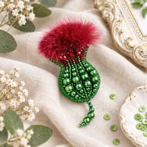 May include: A thistle-shaped brooch crafted with green beads and pearls, featuring a vibrant red feathered top and red crystal accents. The design includes a small beaded stem extending from the base, showcasing intricate beadwork and texture.