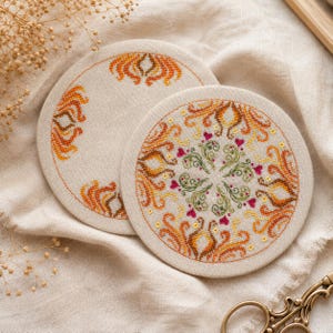 Two embroidered fabric pieces prepared for assembling the dimensional pumpkin pincushion. Decorative cross stitch borders, ornamental motifs, and autumn colors are visible. This image illustrates how the pincushion is constructed from stitched panels before finishing and stuffing.