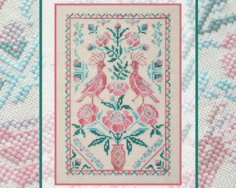 以下が含まれることがあります： 薄いベージュの布地に施されたクロスステッチ刺繍。中央の花瓶から伸びるピンクとティールブルーの花々の両側に、向かい合う2羽の冠羽のある鳥が描かれています。デザインはピンクとティールブルーの幾何学模様の縁取りで囲まれています。