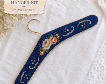 May include: A dark blue clothes hanger featuring a cross-stitch floral pattern with cream and pink roses, green leaves, and gold trim. The hanger has a gold-coloured metal hook. A label above reads: CROSS STITCH HANGER KIT, DIY EMBROIDERY KIT.