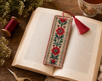 May include: A rectangular fabric bookmark featuring a cross-stitch floral pattern in shades of red, green, and black on a beige background. The bookmark is finished with a decorative red tassel attached to the top edge with a thin red cord.