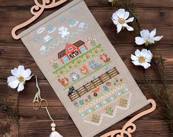 May include: A rectangular linen wall hanging featuring a cross-stitch pattern of a red barn, various cats, garden fences, and flowers. The fabric is held by two decorative light-coloured wooden hangers at the top and bottom, with a tassel attached.