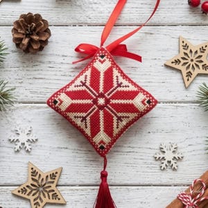 May include: A diamond-shaped cross-stitch ornament featuring a red and cream snowflake pattern. The ornament is suspended by a red satin ribbon and finished with a matching red tassel at the bottom, creating a festive holiday decoration.