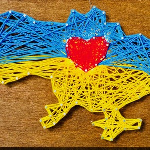 Könnte beinhalten: Ein String-Art-Bild der ukrainischen Karte, hergestellt aus blauen und gelben Fäden, mit einem roten Herzen in der Mitte. Das Kunstwerk ist auf einer Holzoberfläche angebracht und zeigt die Farben der ukrainischen Flagge.