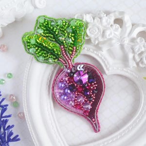 May include: A handcrafted brooch shaped like a beet, featuring vibrant green beaded leaves and a root section embellished with purple, magenta, and dark red sequins, crystals, and pearls. The design includes a central faceted purple crystal.