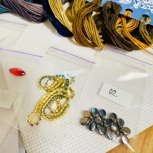 Puede incluir: Un conjunto de suministros de bordado, que incluye madejas de hilo de colores, peque&ntilde;as bolsas de pl&aacute;stico con cuentas y adornos, y una gu&iacute;a de patrones florales azules. La imagen muestra diversos materiales de manualidades para proyectos de costura.