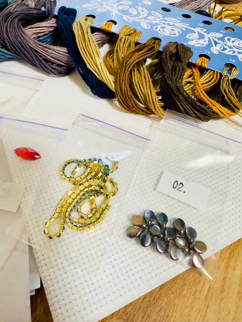 Puede incluir: Un conjunto de suministros de bordado, que incluye madejas de hilo de colores, peque&ntilde;as bolsas de pl&aacute;stico con cuentas y adornos, y una gu&iacute;a de patrones florales azules. La imagen muestra diversos materiales de manualidades para proyectos de costura.