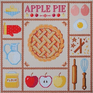 Può includere: Design a punto croce con una torta di mele e ingredienti per la cottura. L'immagine include una torta, uova, mele, farina, burro, un mattarello e una frusta. Le parole "APPLE PIE" sono ricamate.