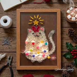 Puede incluir: Una pieza de arte de hilo enmarcada que presenta un gato decorado como un árbol de Navidad. El gato está hecho de hilo blanco con luces de pompón de colores. Un lazo rojo y una estrella adornan la parte superior. Fondo marrón.