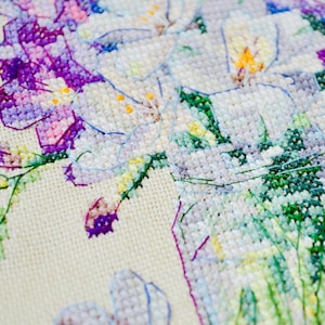 May include: Close-up of a cross-stitch floral design. The embroidery features white, purple, and green flowers with yellow centers. The needlework is on a cream-colored fabric, showcasing the detailed stitches and color variations.