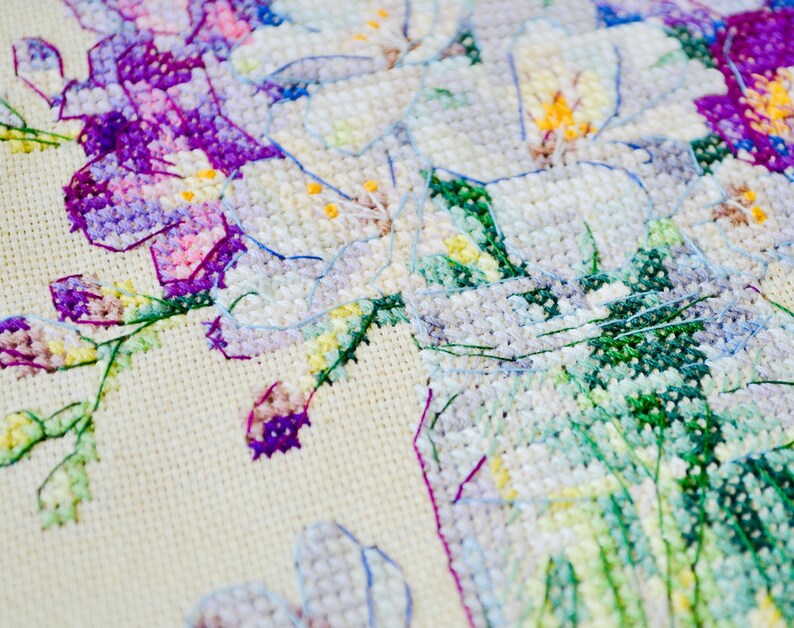 May include: Close-up of a cross-stitch floral design. The embroidery features white, purple, and green flowers with yellow centers. The needlework is on a cream-colored fabric, showcasing the detailed stitches and color variations.
