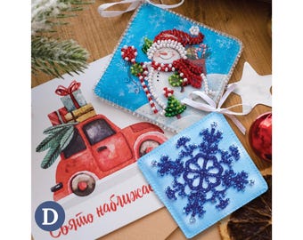 May include: Christmas ornaments featuring a snowman and a snowflake design. The snowman ornament has a red hat and scarf, and the snowflake ornament is blue. A Christmas card with a red truck carrying gifts is also present.