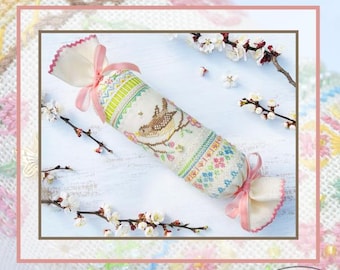 以下が含まれることがあります： ピンクの花が咲く枝にとまった茶色の鳥のクロスステッチ刺繍が施された円筒形の布製サシェ。クリーム色の生地の両端は淡いピンクのリボンで結ばれており、縁はピンクのスカラップ仕上げになっています。