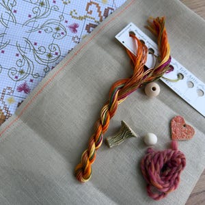 May include: A cross-stitch kit featuring a beige linen fabric, a printed pattern chart, a skein of variegated orange, yellow, and purple embroidery floss, two wooden beads, a small ceramic heart charm, and a textured green ceramic piece.
