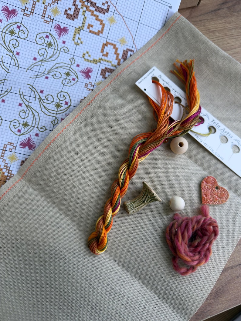 May include: A cross-stitch kit featuring a beige linen fabric, a printed pattern chart, a skein of variegated orange, yellow, and purple embroidery floss, two wooden beads, a small ceramic heart charm, and a textured green ceramic piece.
