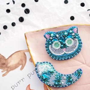 Puede incluir: Dos broches de cuentas hechos a mano con forma de cara de gato y media luna. La cara del gato está detallada con cuentas de color azul verdoso, azul y blanco, ojos verdes y detalles en rosa. La media luna está adornada con cuentas a juego.
