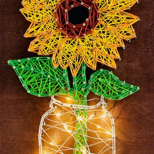 May include: A string art sunflower in a mason jar with fairy lights. The sunflower has yellow and orange petals, a dark red center, and green leaves. The jar and stem are also green. The background is brown.