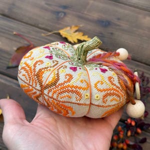 Scale photo showing the finished pumpkin pincushion resting in the palm of a hand. The image helps visualize the compact 11 × 11 cm size while highlighting the dimensional shape, detailed embroidery, ceramic stem, decorative thread, and handcrafted finishing details.