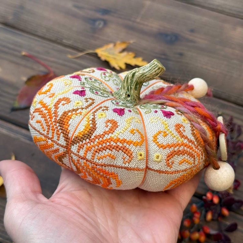 Scale photo showing the finished pumpkin pincushion resting in the palm of a hand. The image helps visualize the compact 11 × 11 cm size while highlighting the dimensional shape, detailed embroidery, ceramic stem, decorative thread, and handcrafted finishing details.