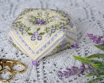 May include: A pentagonal pincushion made of cream-coloured fabric featuring intricate cross-stitch embroidery of small purple and green floral patterns, accented with tiny clear beads and a single purple bead at each corner point.