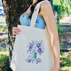 May include: Cream-colored tote bag featuring a watercolor illustration of purple and white flowers in a glass jar. The bag has long, natural-colored straps and is held by a person wearing a blue top. The background includes a tree and greenery.