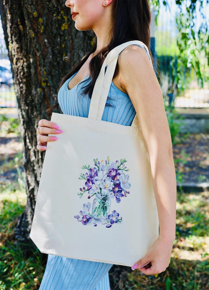 May include: Cream-colored tote bag featuring a watercolor illustration of purple and white flowers in a glass jar. The bag has long, natural-colored straps and is held by a person wearing a blue top. The background includes a tree and greenery.